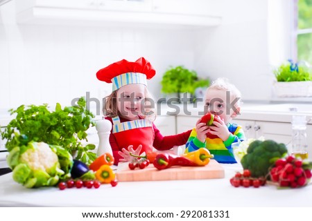 Kids cooking fresh vegetable salad in a white kitchen. Children cook vegetables for vegetarian lunch. Toddler and baby eat healthy dinner. Boy and girl preparing and eating raw meal. Child nutrition.