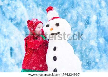 Funny little toddler girl in a red knitted Nordic hat and warm coat playing with a snow. Kids play outdoors in winter. Children having fun at Christmas time. Child building snowman at Xmas.
