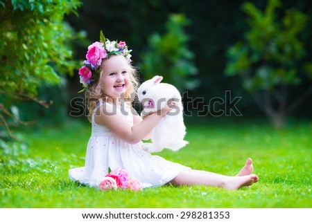 Little girl with real rabbit. Child playing with pet bunny. Kids play with animals. Children at Easter egg hunt. Toddler kid in flower crown and white birthday dress in sunny summer garden.