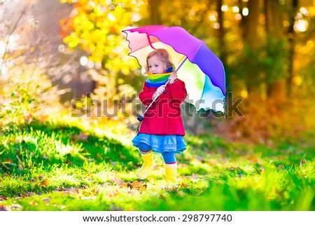 Little girl playing in the rain in autumn park. Child holding umbrella walking in the forest on a sunny fall day. Children playing outdoors with yellow maple leaf. Toddler girl picking golden leaves.