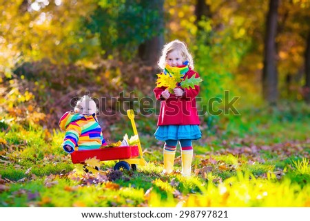 Brother and sister play in autumn park with golden leaves. Baby boy in a wheel barrow. Two kids, boy and girl walk in the forest on a sunny fall day. Children playing outdoors with yellow maple leaf.