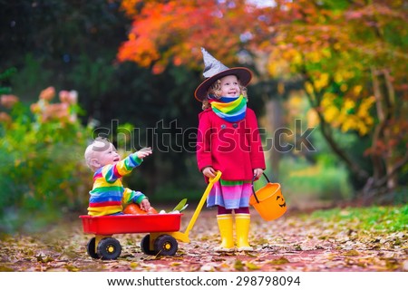 Little girl in witch costume and baby boy in wheel barrow holding a pumpkin playing in autumn park. Kids at Halloween trick or treat. Toddler with jack-o-lantern. Children with candy bucket in forest.