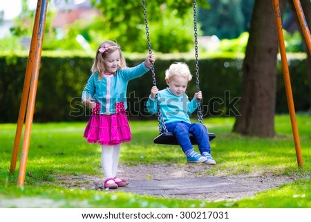 Little boy and girl on a playground. Child playing outdoors in summer. Kids play on school yard. Happy kid in kindergarten or preschool. Children having fun at daycare play ground. Toddler on a swing.