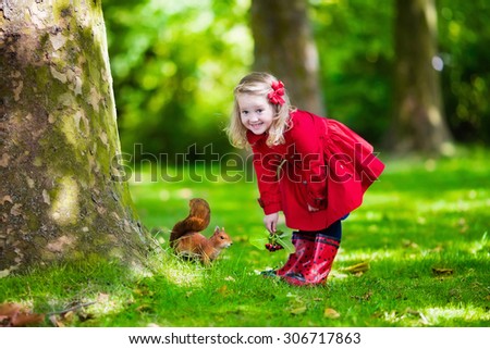 Girl feeding squirrel in autumn park. Little girl in red trench coat and rain boots watching wild animal in fall forest with golden oak and maple leaves. Children play outdoors. Kids playing with pets