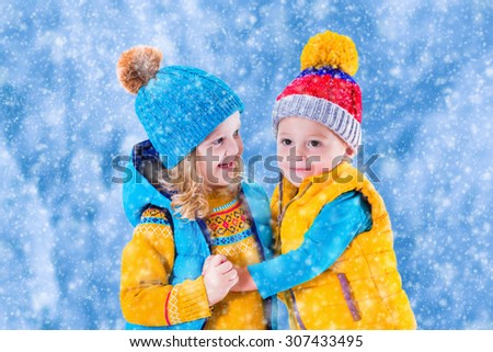 Little girl and boy in yellow and blue knitted hat catching snowflakes in winter park on Christmas eve. Kids play outdoor in snowy winter forest. Children catch snow flake on Xmas. Toddler kid playing