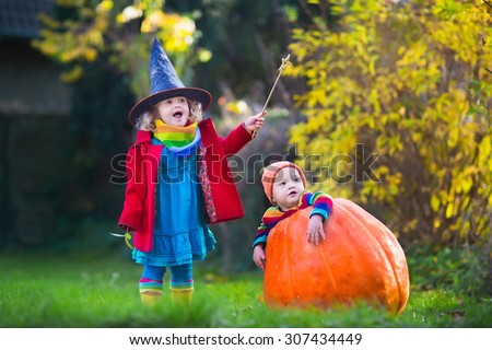 Little girl in witch costume and baby boy in huge pumpkin playing in autumn park. Kids at Halloween trick or treat. Toddler with jack-o-lantern. Children with candy bucket in forest.