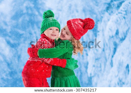 Little girl and boy in red and green knitted hat catching snowflakes in winter park on Christmas eve. Kids play outdoor in snowy winter forest. Children catch snow flakes on Xmas. Toddler kid playing.