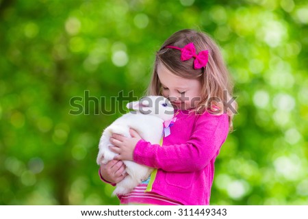 Children play with real rabbit. Laughing child at Easter egg hunt with white pet bunny. Little toddler girl playing with animal in the garden. Summer outdoor fun for kids with pets.