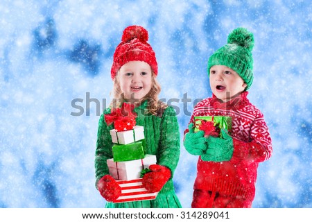 Little girl and boy in red and green knitted hat holding Christmas present boxes in winter park on Xmas eve. Kids play outdoor in snowy winter forest. Children opening presents. Toddler kid with gifts