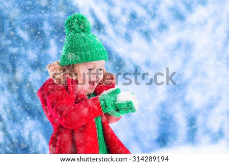 Little girl in red jacket and green knitted hat catching snowflakes in winter park on Christmas eve. Kids play outdoor in snowy winter forest. Children catch snow flakes on Xmas. Toddler kid playing.