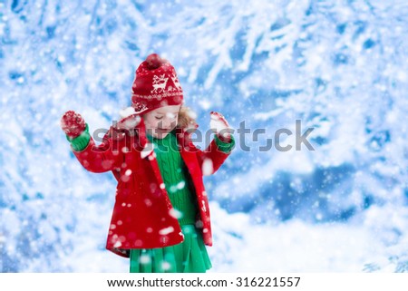 Little girl in red jacket and green knitted dress catching snowflakes in winter park on Christmas eve. Kids play outdoor in snowy winter forest. Children catch snow flakes on Xmas. Toddler kid playing