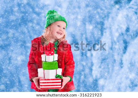 Little girl in red and green knitted hat holding Christmas present boxes in winter park on Xmas eve. Kids play outdoor in snowy winter forest. Children opening presents. Toddler kid playing with gifts