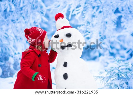 Funny little toddler girl in a red knitted Nordic hat and warm coat playing with a snow man. Kids play outdoors in winter. Children having fun at Christmas time. Child building snowman at Xmas.
