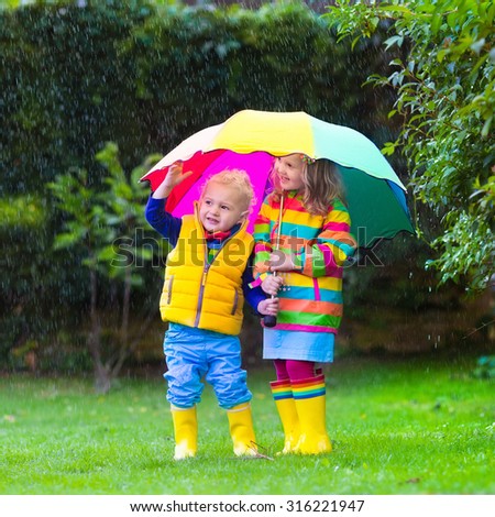 Little girl with colorful umbrella playing in the rain. Kids play outdoors by rainy weather in fall. Autumn fun for children. Toddler kid in raincoat and boots walking in the garden. Summer shower.