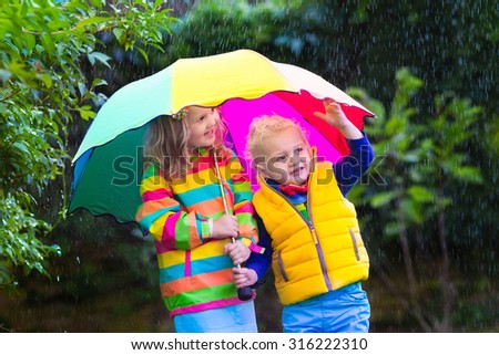 Little girl and boy with colorful umbrella playing in the rain. Kids play outdoor by rainy weather in fall. Autumn fun for children. Toddler kid in raincoat and boots walk in the garden. Summer shower