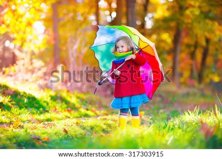 Little girl playing in the rain in autumn park. Child holding umbrella walking in the forest on a sunny fall day. Children playing outdoors with yellow maple leaf. Toddler girl picking golden leaves