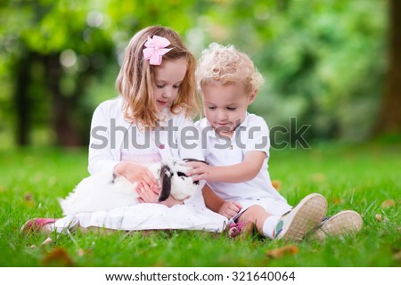 Children play with real rabbit. Brother and sister at Easter egg hunt with white pet bunny. Little baby boy and toddler girl playing with animal in the garden. Summer outdoor fun for kids with pets.