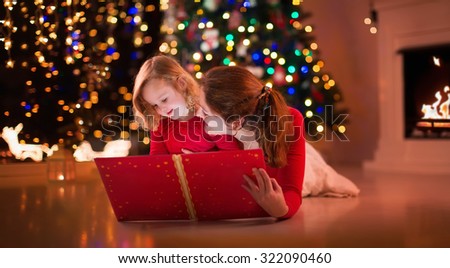 Mother and daughter read a book at fireplace on Christmas eve. Family with child celebrating Xmas. Decorated living room with tree, fire place and candles. Winter evening at home for parents and kids.