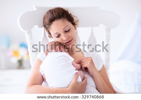 Young mother holding her newborn child. Mom nursing baby. Woman and new born boy relax in a white bedroom with rocking chair and blue crib. Nursery interior. Mother breast feeding baby. Family at home