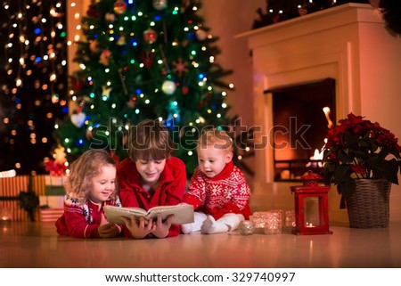 Children read a book and open gifts at fireplace on Christmas eve. Family with child celebrating Xmas. Decorated living room with tree, fire place, candles. Winter evening at home for parents and kids