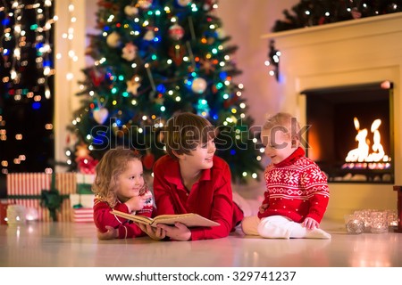 Children read a book and open gifts at fireplace on Christmas eve. Family with child celebrating Xmas. Decorated living room with tree, fire place, candles. Winter evening at home for parents and kids