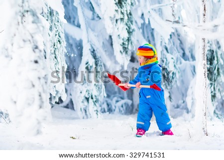 Little girl shoveling snow on home drive way. Beautiful snowy garden or front yard. Child with shovel playing outdoors in winter season. Family removing snow after blizzard. Kids play outside.