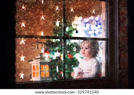 Little girl watching snow through window. Family with children on Christmas eve in decorated living room with tree and candles. Kids open Xmas presents. Cozy winter evening at home. View from outside.