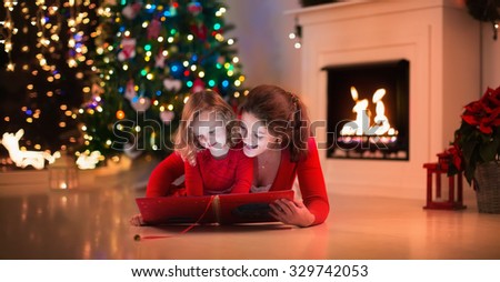 Mother and daughter read a book at fireplace on Christmas eve. Family with child celebrating Xmas. Decorated living room with tree, fire place and candles. Winter evening at home for parents and kids.