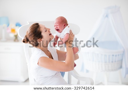 Young mother holding her newborn child. Mother comforting crying hungry baby. Woman and new born boy relax in a white bedroom with rocking chair and blue crib. Nursery interior. Family at home.