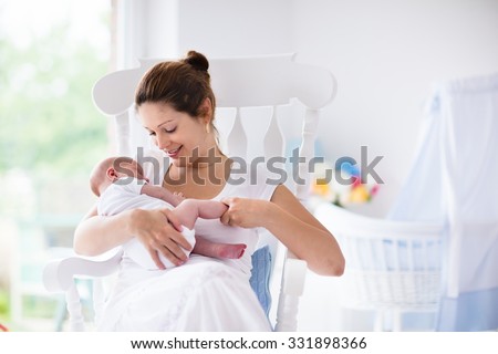 Young mother holding her newborn child. Mom nursing baby. Woman and new born boy relax in a white bedroom with rocking chair and blue crib. Nursery interior. Mother breast feeding baby. Family at home