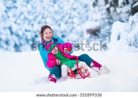 Young mother and little girl enjoying sleigh ride. Child sledding. Toddler kid riding sledge. Children play outdoors in snow. Kids sled in snowy park. Outdoor winter fun for family Christmas vacation.