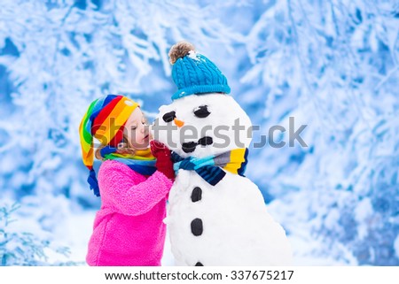 Funny little toddler girl in a colorful hat and warm coat playing with a snow man. Kids play outdoors in winter. Children having fun at Christmas time. Child building snowman at Xmas.