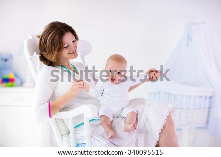 Young mother holding her newborn child. Mom nursing baby. Woman and new born boy in white bedroom with rocking chair and blue crib. Nursery interior. Mother playing with laughing kid. Family at home