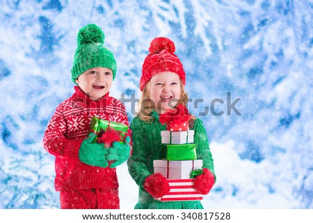 Little girl and boy in red and green knitted hat holding Christmas present boxes in winter park on Xmas eve. Kids play outdoor in snowy winter forest. Children opening presents. Toddler kid with gifts