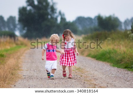 Girl and boy playing in the rain. Kids play outdoor by rainy weather in fall. Autumn fun for children. Toddler kid and baby walk in the garden. Summer shower. Brother and sister run holding hands.