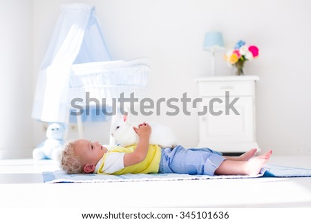 Cute blond curly baby boy playing with rabbit in a white sunny bedroom. Kids and pets at home. Children and animals play indoors. Funny toddler kid holding Easter bunny. Child taking care of an animal