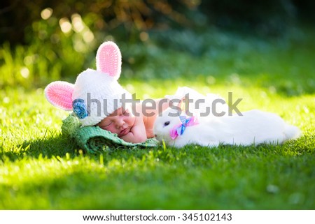 Cute newborn baby boy in bunny costume wearing hat with ears and white diaper and a real rabbit in sunny green garden during Easter egg hunt. Children and pets friendship. Kids and animals.