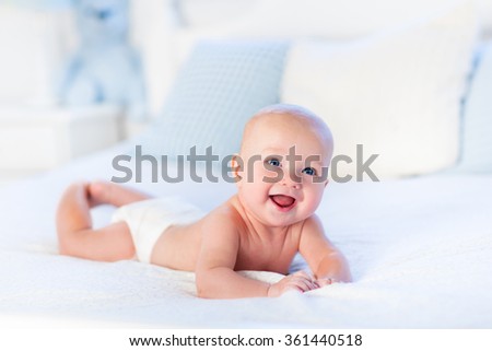 Baby boy wearing diaper in white sunny bedroom. Newborn child relaxing in bed. Nursery for children. Textile and bedding for kids. Family morning at home. New born kid during tummy time with toy bear.