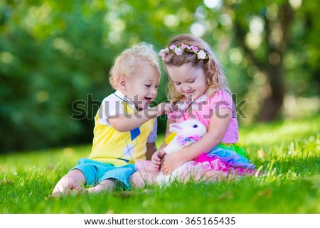 Children play with real rabbit. Brother and sister at Easter egg hunt with white pet bunny. Little baby boy and toddler girl playing with animal in the garden. Summer outdoor fun for kids with pets.