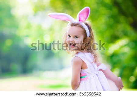 Little girl having fun on Easter egg hunt. Kids in bunny ears and rabbit costume. Children searching for eggs in the garden. Toddler kid playing outdoor. Child laughing and smiling on a spring day