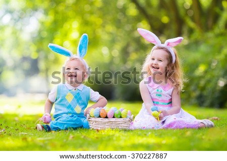 Little boy and girl having fun on Easter egg hunt. Kids in bunny ears and rabbit costume. Children with colorful eggs in a basket. Toddler kid and baby play outdoor on sunny spring day. Family holiday