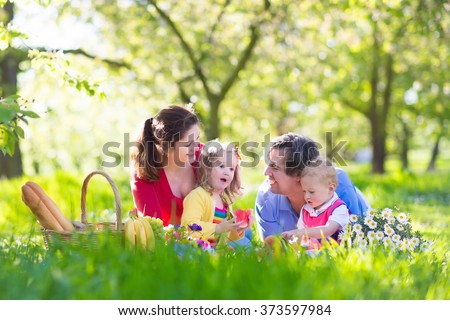 Family with children enjoying picnic in spring garden. Parents and kids having fun eating lunch outdoors in summer park. Mother, father, son and daughter eat fruit and sandwiches on colorful blanket.