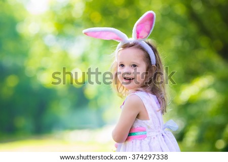 Little girl having fun on Easter egg hunt. Kids in bunny ears and rabbit costume. Children with colorful eggs in a basket. Toddler kid playing outdoor. Child holding blank white board for your text.