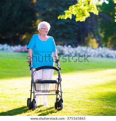 Happy senior handicapped lady with a walking disability enjoying a walk in a sunny park pushing her walker or wheel chair, aid and support during retirement concept.