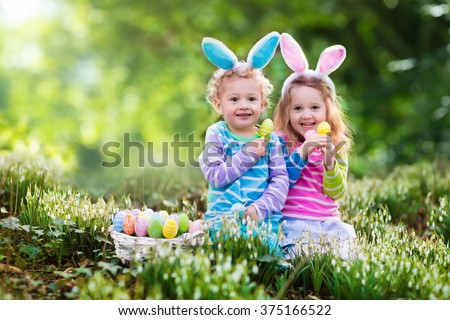 Kids on Easter egg hunt in blooming spring garden. Children with bunny ears searching for colorful eggs in snow drop flower meadow. Toddler boy and preschooler girl in rabbit costume play outdoors.