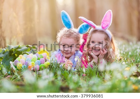 Kids on Easter egg hunt in blooming spring garden. Children with bunny ears searching for colorful eggs in snow drop flower meadow. Toddler boy and preschooler girl in rabbit costume play outdoors.