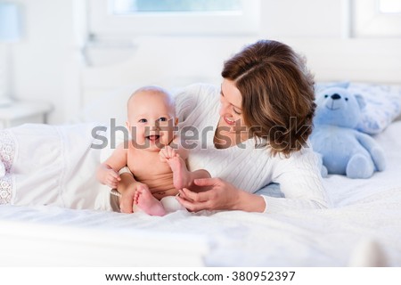 Mother and child on a white bed. Mom and baby boy in diaper playing in sunny bedroom. Parent and little kid relaxing at home. Family having fun together. Bedding and textile for infant nursery.
