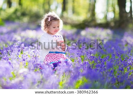 Little girl playing in sunny blooming garden. Baby on Easter egg hunt in blue bell flower meadow. Toddler child picking bluebell flowers. Kids play outdoors. Spring fun for family with children.
