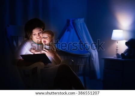 Mother and baby reading a book in dark bedroom. Mom and child read books before bed time. Family in the evening. Kids room interior with night lamp and bassinet. Parent holding infant next to crib.