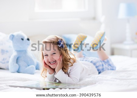 Funny happy toddler girl reading a book and playing with her toy teddy bear in bed. Kids play at home. White nursery. Child in sunny bedroom. Children read and study. Interior for baby and young kid.